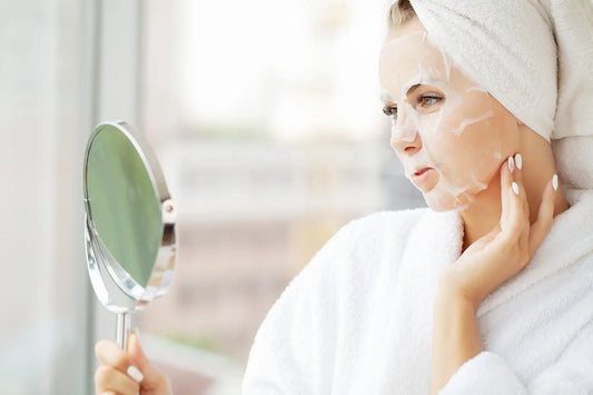 Woman applying sheet mask while looking in mirror wearing towel